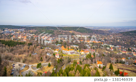 A wide view of a small town surrounded by trees and hills. Several buildings are visible, with roads and cars parked nearby. The scene shows a clear sky above. A wide view of a small town surrounded by trees and hills. Several buildings are visible, with roads and cars parked nearby. The scene shows a clear sky above. 138146939
