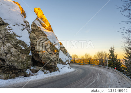 A winding road wraps around a large rock covered in snow. The sun sets in the background, casting light on the rock. Trees stand bare around the road during winter. A winding road wraps around a large rock covered in snow. The sun sets in the background, casting light on the rock. Trees stand bare around the road during winter. 138146942