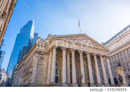 The Royal Exchange in London stands among modern structures on a clear day. People walk nearby, and the British flag flies high above the historic site. 138146955