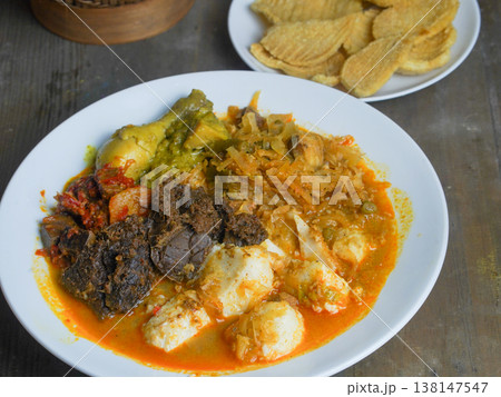 Traditional Indonesian ketupat sayur dish served on a white plate with a wooden container and food items in the background on a rustic table. 138147547