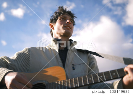 The wild and free spirit of the guitarist. A young guy plays the guitar against a background of blue sky and white clouds, a view from below. An inspired musician composes a new melody on the guitar 138149685
