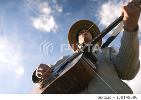 A young man in a cowboy hat plays guitar against a background of blue sky and white clouds, a view from below. Cowboy gives guitar lessons. The rebellious spirit A young man in a cowboy hat plays guitar against a background of blue sky and white clouds, a view from below. Cowboy gives guitar lessons. The rebellious spirit 138149686
