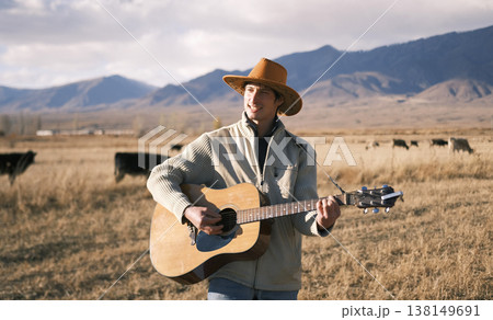 A handsome man in a cowboy hat sings songs with a guitar. A musician plays the guitar in a farmer's field. The sky is overcast, and mountains and a herd of cows 138149691