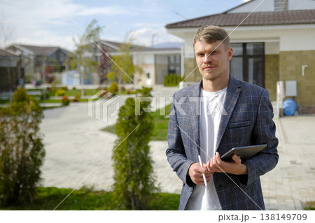 A real estate insurer. Portrait of a realtor or sales manager. A confident, successful, handsome man in a suit with a tablet and a stylus in his hands stands in front of a country house A real estate insurer. Portrait of a realtor or sales manager. A confident, successful, handsome man in a suit with a tablet and a stylus in his hands stands in front of a country house 138149709