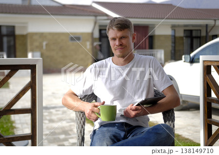 A young successful freelancer is sitting on the veranda of his country house with a tablet and a cup of coffee in his hands. The insurer. The realtor is sitting on a chair 138149710