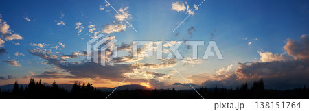 Serene evening sky with hues of blue and soft pink clouds as sun sets. Horizon glows gently, casting light over silhouetted pine trees and rolling hills, creating peaceful and expansive landscape. 138151764