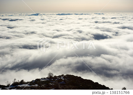 Snow-covered mountain slope with scattered rocks and trees. Landscape bathed in soft sunlight, while dense layer of clouds stretches out below, creating dramatic and ethereal scene. 138151766