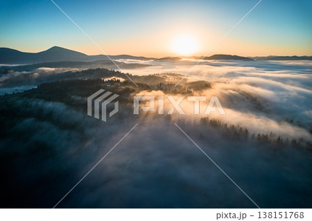 Stunning aerial view of sunrise over mountainous landscape, with thick fog blanketing valleys and hills. Sun's golden rays pierce through mist, casting serene glow over the entire scene. Stunning aerial view of sunrise over mountainous landscape, with thick fog blanketing valleys and hills. Sun's golden rays pierce through mist, casting serene glow over the entire scene. 138151768