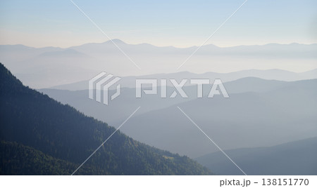 Misty morning in mountains with layers of forested hills fading into distance. Rising sun casts golden light on green slope, creating serene and peaceful landscape. 138151770