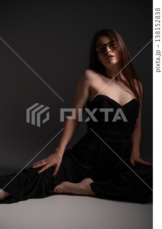 Young woman in stylish black strapless jumpsuit with glasses posing under dramatic lighting, sitting on the floor with confidence and elegance 138152838