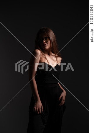 Elegant young woman wearing black strapless dress and stylish yellow tint glasses posing in dark studio, expressing confidence, fashion, and mystery Elegant young woman wearing black strapless dress and stylish yellow tint glasses posing in dark studio, expressing confidence, fashion, and mystery 138152845