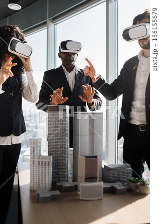 Three professionals in business attire wearing VR headsets interact with a miniature city model, exploring architectural designs in a virtual reality environment 138155679