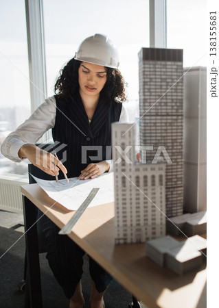 A female architect wearing a hard hat reviews blueprints and building models at her desk with a large window in the background A female architect wearing a hard hat reviews blueprints and building models at her desk with a large window in the background 138155681