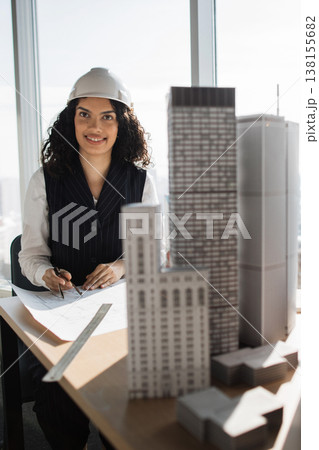A female architect wearing a hard hat reviews blueprints and works with miniature building models in an office with a city view A female architect wearing a hard hat reviews blueprints and works with miniature building models in an office with a city view 138155682