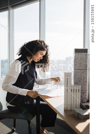 A designer studies scale models and technical drawings by a bright window in a high-rise office overlooking the city skyline A designer studies scale models and technical drawings by a bright window in a high-rise office overlooking the city skyline 138155691