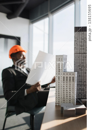 An architect wearing an orange hard hat reviews blueprints while sitting in an office with architectural models on a desk 138155695