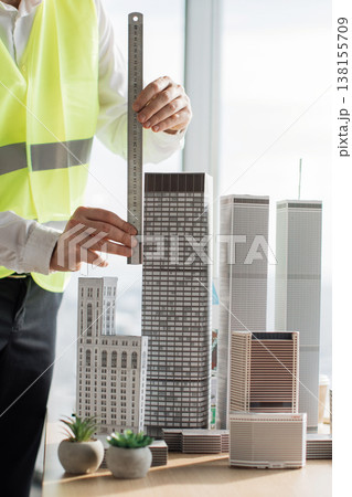 An architect wearing a safety vest uses a ruler to measure a detailed model of a skyscraper, surrounded by other architectural models 138155709
