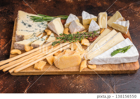 Brie Camembert Parmesan and Roquefort arranged with nuts herbs and breadsticks on a rustic wooden board over a dark textured background. Brie Camembert Parmesan and Roquefort arranged with nuts herbs and breadsticks on a rustic wooden board over a dark textured background. 138156550