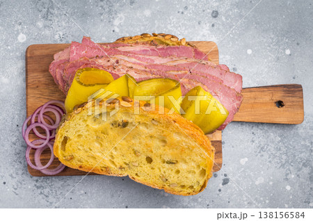 Stacked sliced pastrami in seed crust bread topped with pickles, served on a rustic wood board against gray stone surface. Stacked sliced pastrami in seed crust bread topped with pickles, served on a rustic wood board against gray stone surface. 138156584