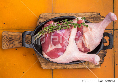 Raw duck legs in a black cast iron skillet, garnished with fresh thyme, resting on a rustic wooden cutting board against an orange tiled background. Raw duck legs in a black cast iron skillet, garnished with fresh thyme, resting on a rustic wooden cutting board against an orange tiled background. 138156648