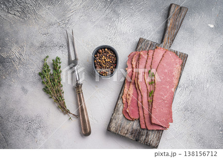 Thin pastrami slices with thyme on a rustic wooden serving board, placed over a textured gray background. Smoked deli beef ready for sandwiches or recipes. 138156712