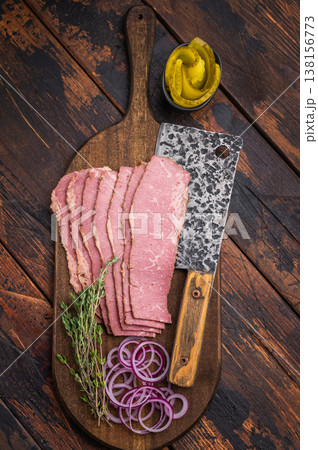 Thinly sliced pastrami on a wooden cutting board with a hammered knife, red onion rings and thyme sprigs on a dark textured wood background. Smoked deli beef ready for meals. 138156773