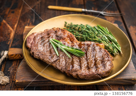 Dry aged grilled striploin steak grilled to perfection on a golden plate with rosemary and thyme on rustic wooden background Dry aged grilled striploin steak grilled to perfection on a golden plate with rosemary and thyme on rustic wooden background 138156776