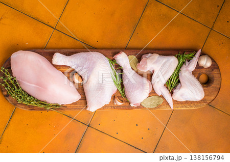 Fresh assortment of chicken breast, wings, drumsticks, and leg quarters on a wooden cutting board with spices, set on an orange kitchen counter. 138156794