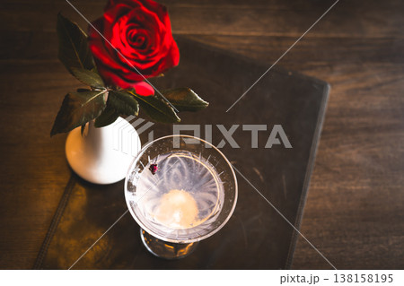 Top view of a crystal cocktail glass and a red rose in a white vase on a dark wooden table. Moody vintage still life with soft lighting and film grain. Ideal for romantic and celebration designs. 138158195