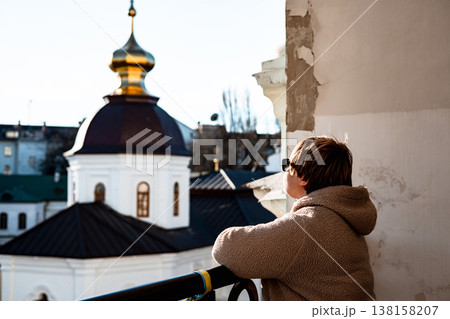 Candid shot of a person in a brown jacket, sunglasses standing on a balcony, admiring the view of a traditional orthodox church with a gold dome and white walls. A moment of historical exploration. 138158207
