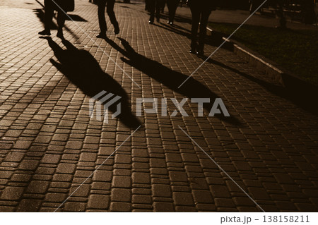 Candid, high-contrast shot of people's silhouettes and long shadows walking on a paved urban sidewalk. The dramatic lighting creates an atmospheric effect, capturing a moment of everyday city life. 138158211