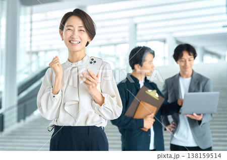 スマートフォンを手にガッツポーズで喜ぶ笑顔の日本人ビジネス女性と背景のチーム 138159514