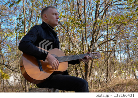 Blonde man playing string guitar outdoors in autumn forest. Concept of sound therapy, mental health and wellness rituals. Calmness tranquility audio-sensory practices. Aura farming energy Blonde man playing string guitar outdoors in autumn forest. Concept of sound therapy, mental health and wellness rituals. Calmness tranquility audio-sensory practices. Aura farming energy 138160431