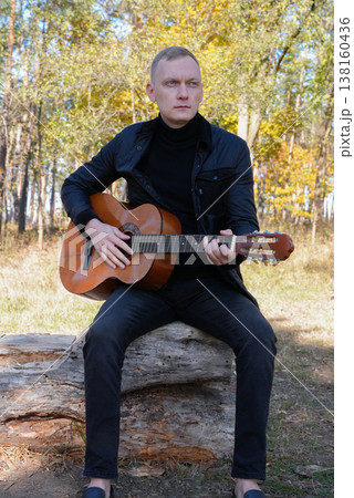 Portrait of smiling caucasian artist plays wooden acoustic guitar in autumnal park. Young songwriter plays string musical instrument hobby outside in nature fall time. Audio music healing Portrait of smiling caucasian artist plays wooden acoustic guitar in autumnal park. Young songwriter plays string musical instrument hobby outside in nature fall time. Audio music healing 138160436