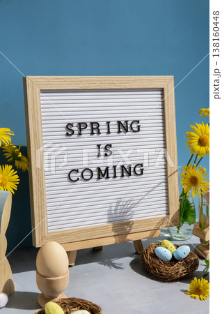 Phrase SPRING IS COMING traditional greeting to christian holiday. Festive mood composition with bunnies and yellow flowers on pastel blue background. Sustainable natural materials Phrase SPRING IS COMING traditional greeting to christian holiday. Festive mood composition with bunnies and yellow flowers on pastel blue background. Sustainable natural materials 138160448