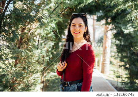 Smiling slavic appearance young woman walking in a zoo park wearing red sweater and jeans surrounded by green trees in warm sunlight looking at camera 138160748