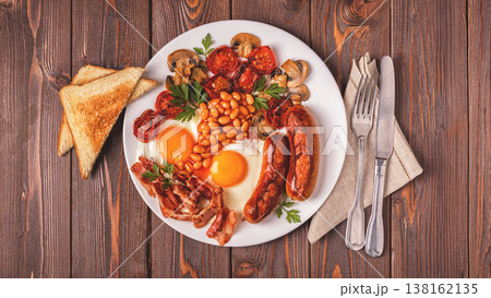 Traditional full English breakfast with fried eggs, sausages, beans, mushrooms, grilled tomatoes and bacon on wooden background. Traditional full English breakfast with fried eggs, sausages, beans, mushrooms, grilled tomatoes and bacon on wooden background. 138162135