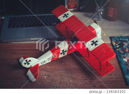 Horizontal view of a handcrafted red and white wooden triplane model with a propeller, resting on a wooden workbench next to a laptop in a workshop setting. 138163101