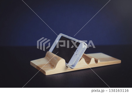 Horizontal close-up of a handcrafted light wood phone stand with a carved wave surface, securely holding a white smartphone at an angle against a dark blue background. 138163116