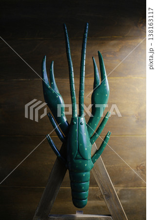 Vertical front view of a large handcrafted wooden crayfish sculpture painted dark green with light green spots, displayed on a wooden easel against a plank wall. 138163117
