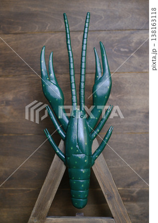 Vertical front view of a handcrafted wooden crayfish sculpture painted dark green with light green spots, displayed on a wooden easel against a plank wall. Vertical front view of a handcrafted wooden crayfish sculpture painted dark green with light green spots, displayed on a wooden easel against a plank wall. 138163118
