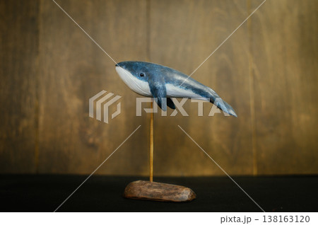 Horizontal side view of a handcrafted wooden blue whale sculpture, featuring a white underbelly and displayed on a thin vertical stand with a rustic base. 138163120