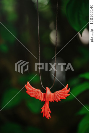 Vertical view of a handcrafted vibrant red wooden phoenix pendant with spread wings, suspended on a dark cord against a lush green forest background 138163140