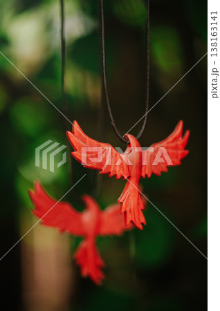 Vertical artistic view of two handcrafted red wooden phoenix pendants, with one in sharp focus and another blurred in the background, against green leaves. 138163141