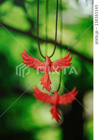 Vertical artistic view of two handcrafted red wooden phoenix pendants, with one in sharp focus and another blurred in the background, against green leaves. 138163143