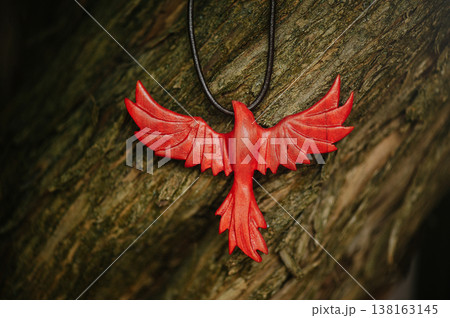 Vertical close-up of a handcrafted red wooden phoenix pendant with spread wings, hanging against the textured bark of a tree. 138163145