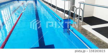 A clean indoor swimming pool features vibrant blue water and red lane dividers. Chrome handrails lead into the water along the stone edge, while black anti-slip mats line the tiled walkway for safety A clean indoor swimming pool features vibrant blue water and red lane dividers. Chrome handrails lead into the water along the stone edge, while black anti-slip mats line the tiled walkway for safety 138163815