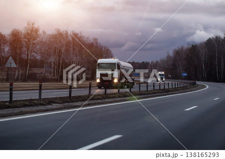Tanker truck with illuminated headlights travels on curved highway through barren landscape under cloudy evening sky, another truck in distance, evening sunset. Copy space for text Tanker truck with illuminated headlights travels on curved highway through barren landscape under cloudy evening sky, another truck in distance, evening sunset. Copy space for text 138165293