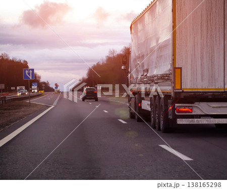Semi-truck with white trailer traveling on highway at dusk during overcast day, freight transportation along road infrastructure, evening sunset. Copy space for text Semi-truck with white trailer traveling on highway at dusk during overcast day, freight transportation along road infrastructure, evening sunset. Copy space for text 138165298