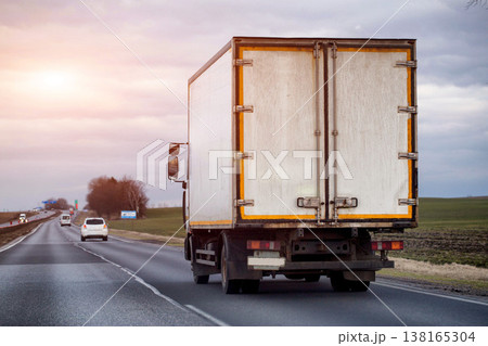 Rear view of cargo truck driving on highway under overcast sky, transportation of goods across landscape, commercial logistics and delivery service, evening sunset. Copy space for text Rear view of cargo truck driving on highway under overcast sky, transportation of goods across landscape, commercial logistics and delivery service, evening sunset. Copy space for text 138165304